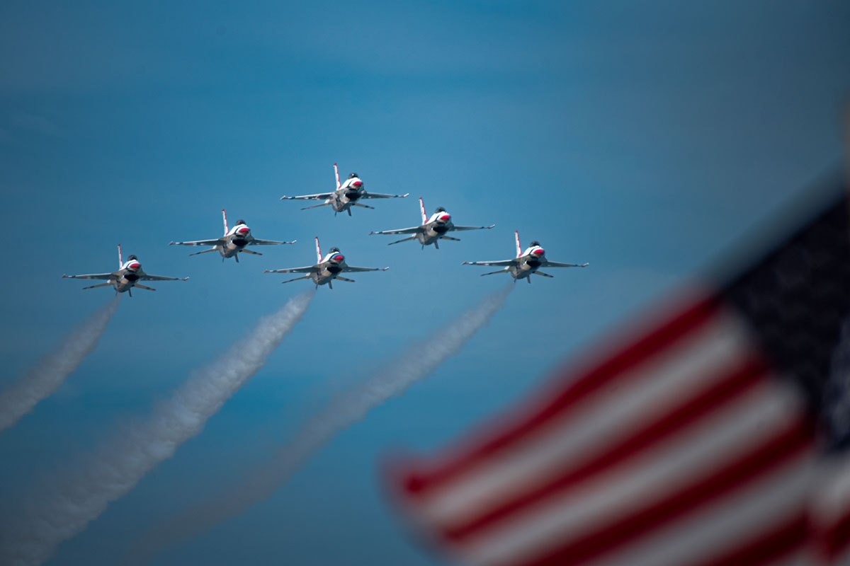 All six jets in formation coming down the beach with an american flag in the foreground
