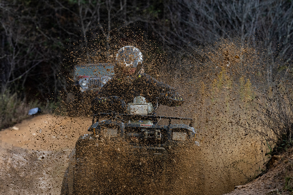 ATV plowing through a mud pit, mud flying all around, a jeep is in the background coming up the trail